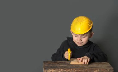Çocuk müteahhitçilik oynuyor. Kask takıyor ve elinde oyuncak bir testereyle tahtayı testereyle kesiyor. koyu arkaplan, boşluğu kopyala