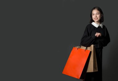 Genç bir esmer kız, gri bir arka planda elinde kağıt torbalarla siyah elbiseler içinde duruyor.