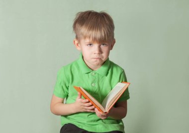 Yeşil tişörtlü, üzgün yüzlü sarışın bir çocuk elinde kitapla oturuyor. yeşil arkaplan