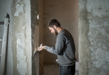 Sakallı bir erkek alçı beton bir duvarı spatulayla kaplıyor.