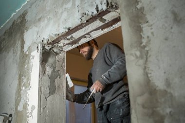 Sakallı bir adam, kapısında delik olan beton bir köşeyi spatülle kaplıyor.