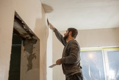 Sakallı bir erkek alçı beton bir duvarı spatulayla kaplıyor.