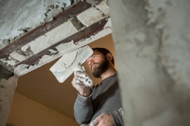 Sakallı bir adam, kapısında delik olan beton bir köşeyi spatülle kaplıyor.