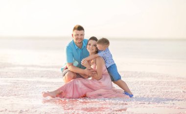 Bir adam, bir kadın ve oğulları pembe bir gölün kıyısında oturuyor, sarılıyor ve eğleniyorlar.