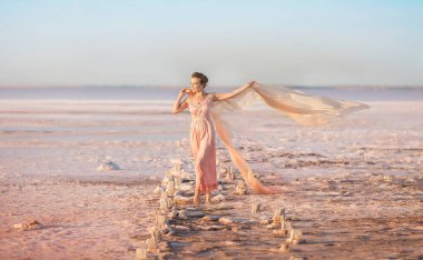 Pembe bir gölün üzerinde pembe bir elbiseyle uçan bir eşarpla duran güzel bir genç kız.