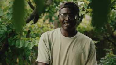Tişörtlü Afro-Amerikalı adam gülümsüyor. Bahçedeki yemyeşil ağaçların önünde duran gözlüklü olgun bir adamın portresi.