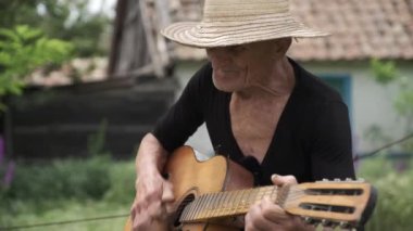 Hasır şapkalı yaşlı adam bulanık arka planda gitar çalıp şarkı söylüyor. Müzikal enstrümanlı emekli köyün bahçesinde yavaş çekimde oturuyor.
