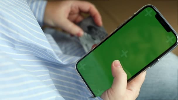 Молодая женщина, девушка в светло-голубой одежде, держит смартфон с зеленым экраном, банковскую карту и делает онлайн покупки через приложение, онлайн денежный перевод — стоковое видео