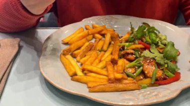 Kadın bir kafede oturur ve patates kızartması ve sebzeleri çatalla yer, tabağı da yakın çekimde. Akşam keyfi.