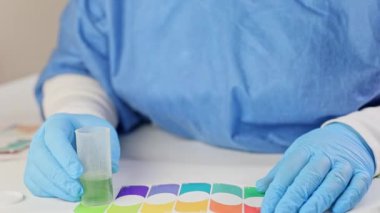 Tıbbi giysi ve eldiven giyen bir laboratuvar asistanı renk ölçeğinde bir test suyu örneğini, kalite ve kimyasal bileşimi kontrol etmek için su testi sürecini kontrol eder.