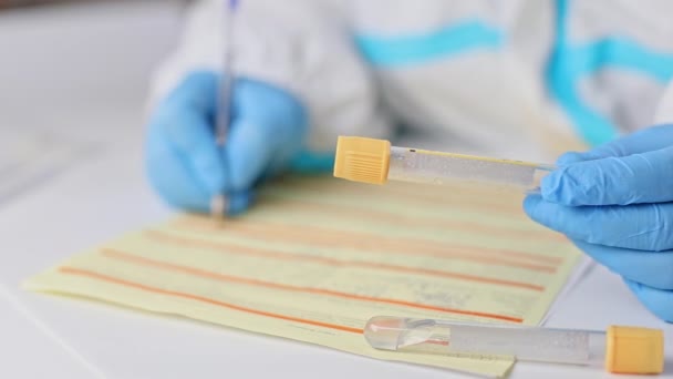 Врач, медсестра или медицинский технолог в медицинских перчатках держит пробирку для анализа крови и пишет, заполняет формы для тестирования пациента на ковид 19, коронавирус. Протокол ПЦР ДНК — стоковое видео