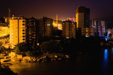 Tipik küçük bir Türk kasabasının gece aydınlığı manzarası. Türkiye 'nin Mersin kenti yakınlarındaki Akdeniz kıyısındaki yerleşim yeri. Binalar arasındaki caminin yüksek minaresi.