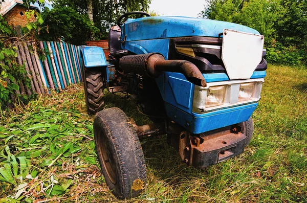 Close-up view of old blue mini tractor on green grass. Mini tractor on old worn tires. A small old mini tractor near rural house. Concept of country vehicle.