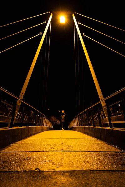 Symmetrical night scene of a cable-stayed bridge with cables forming guide lines to a girl with her back turned walking. The scene is illuminated by a streetlight casting a yellowish light. — Stock Photo, Image
