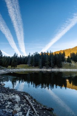 Sonbaharın başlarında ve günbatımından hemen önce İsviçre Alplerinde küçük bir alp gölü. Su sakin ve diğer taraftaki köknar ormanını yansıtıyor. Gökyüzünde sadece üç uçak izi var.