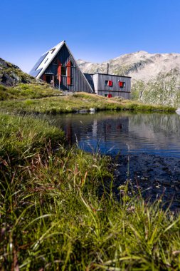 View of the Scaletta hut near the Greina Pass in Blenio, Switzerland. In the foreground is a pond reflecting the hut and the high mountains in the background. The sky is clear.