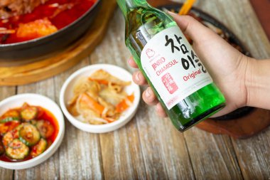 Los Angeles, Kaliforniya, ABD - 08-10-2021: Bir şişe Jinro Chamisul soju tutan bir elin görüntüsü, banchan ve Kore güvecinin üzerinde.