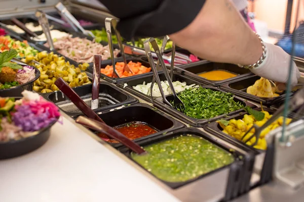 A view of a restaurant food display, featuring a variety of customized topping ingredients. An employee prepares a bowl order.