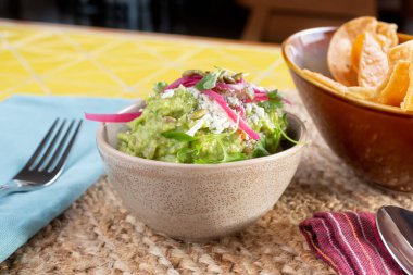 Bir kase dekore edilmiş guacamole çerçevede, mısır tortilla cipsi ile birlikte bir restoran veya mutfak ortamında.