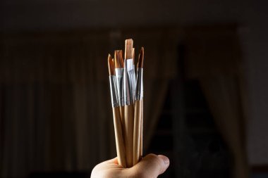 Koyu bir arkaplana karşı boya fırçası koleksiyonu tutan bir elin görüntüsü.