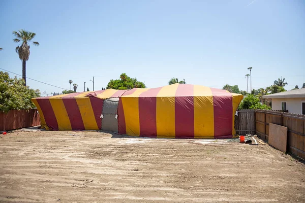 A view of a real estate property covered by a yellow and red striped fumigation tent, seen from the backyard.