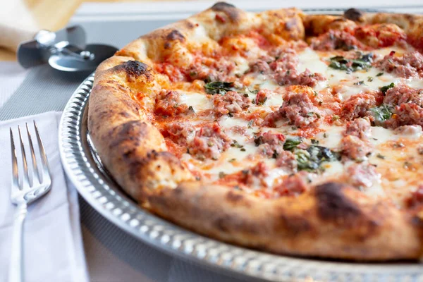 Bir restoranda ya da mutfak ortamında sosis ve otlar gibi malzemelerin yer aldığı rustik bir pizzanın yakın plan görüntüsü.