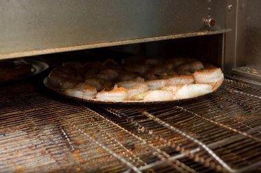 Lokanta mutfağında konveyör fırınında hareket eden bir tepsi dolusu baharatlı karidesin görüntüsü..