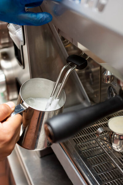 A view of a hand holding a stainless steel pitcher full of steamed milk, next to an espresso machine, in a cafe setting.