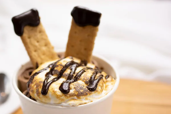 A closeup view of a dessert cup, featuring rolled ice, graham crackers and melted marshmallow topping.