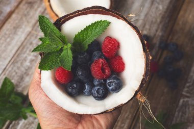 Nane ve böğürtlen dolu yarım hindistan cevizini ahşap bir yüzey masasının üzerinde tutan bir elin yukarıdan görünüşü..