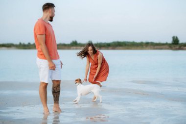 Boş kumsalda, turuncu elbiseli, köpekli genç bir çift.