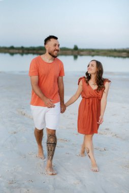 Boş kumsalda, turuncu elbiseli, köpekli genç bir çift.