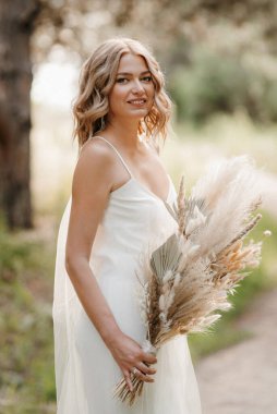 Orman yolunda bir buket kurumuş çiçekle beyaz bir elbise içindeki mutlu gelin kız.