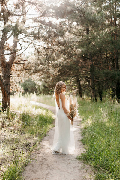 Happy bride girl in a white light dress with a bouquet of dried flowers on a forest path