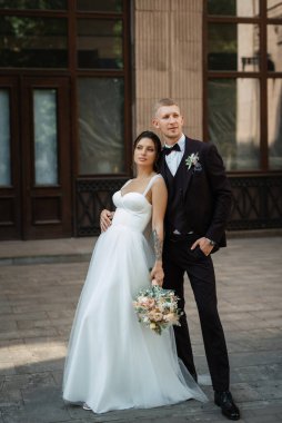 Kahverengi takım elbiseli damat ve şehir ortamında beyaz elbiseli gelin.