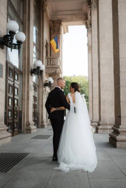 Kahverengi takım elbiseli damat ve şehir ortamında beyaz elbiseli gelin.