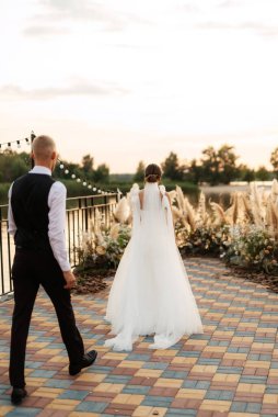 Gelin ve damat, nehrin kıyısındaki bir iskelede sarı bir günbatımının arkaplanına karşı.