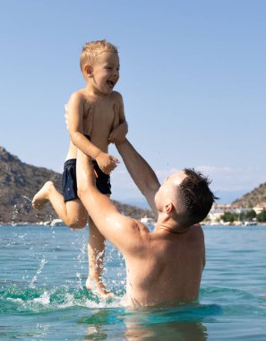 Baba ve oğul denizde oynuyorlar. Baba, oğlunu fırlatıp atıyor. Yunan adaları, su sıçramaları, tatiller, mavi gökyüzü ve güneşli bir gün. Yüksek kalite fotoğraf