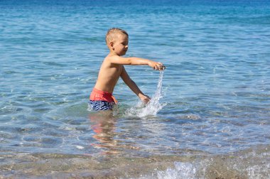 Küçük çocuk denizde oynuyor ve suya taş atıyor.