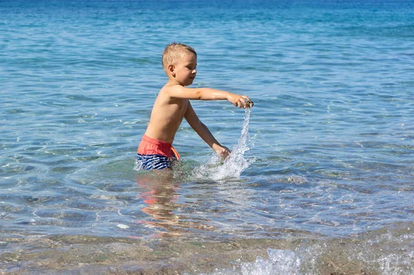 Küçük çocuk denizde oynuyor ve suya taş atıyor.