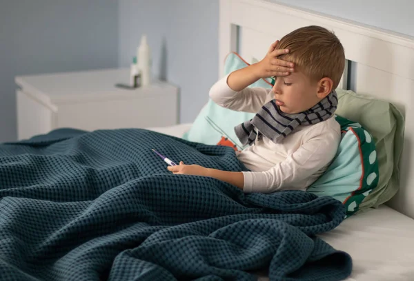 stock image A little boy in a scarf is sick and lies in bed, holding a thermometer in his hands. Seasonal cold concept