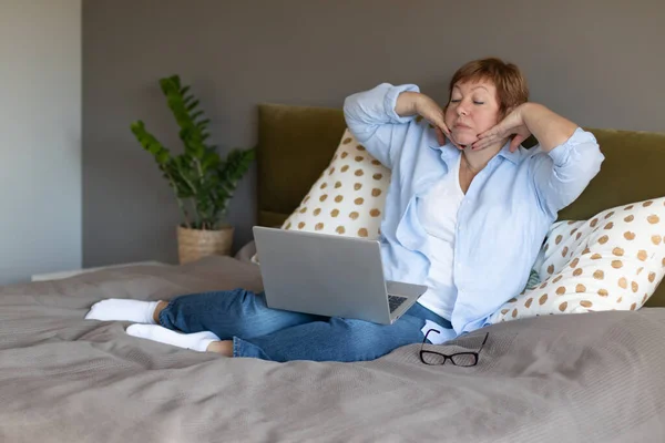 An elderly woman is doing face fitness using video lessons in front of a laptop. Modern technologies and aged people concept