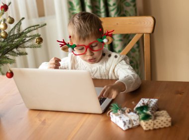  Beyaz kazaklı ve komik gözlükleri olan küçük bir çocuk dizüstü bilgisayarda oturuyor ve Noel Baba 'ya bir mektup yazıyor..