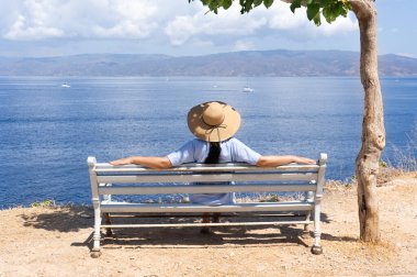 Mavi elbiseli ve büyük hasır şapkalı genç bir kadın sırtıyla bankta oturur ve denize bakar. 