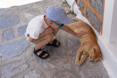 Küçük çocuk sokakta bir kediyi okşuyor.