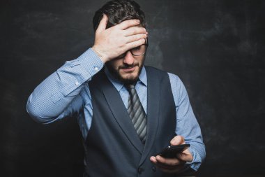 Takım elbiseli bir adam gri arka planda şokta telefona bakar.