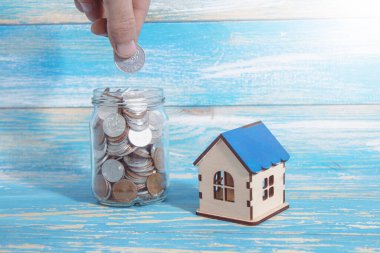 on the table a bank with coins and a house. hand puts a coin