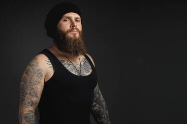 Siyah tişörtlü ve şapkalı, kameraya bakan yakışıklı sakallı bir adamın portresi..