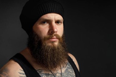 Siyah tişörtlü ve şapkalı, kameraya bakan yakışıklı sakallı bir adamın portresi..