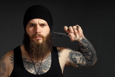 Sakallı cesur bir adamın portresi. Sakalının yanında makası tutuyor ve kameraya bakıyor..